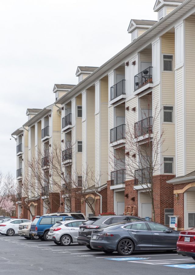 Four-story apartment building with balconies, parked cars in front, and leafless trees on a cloudy day.