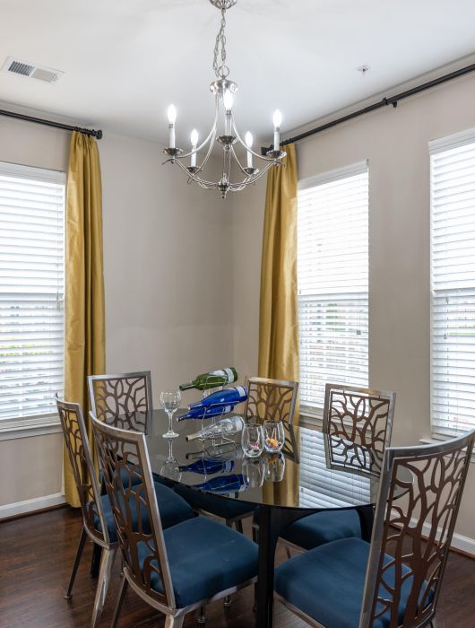 Modern dining room with glass table, four chairs, gold curtains, chandelier, and floor lamp.
