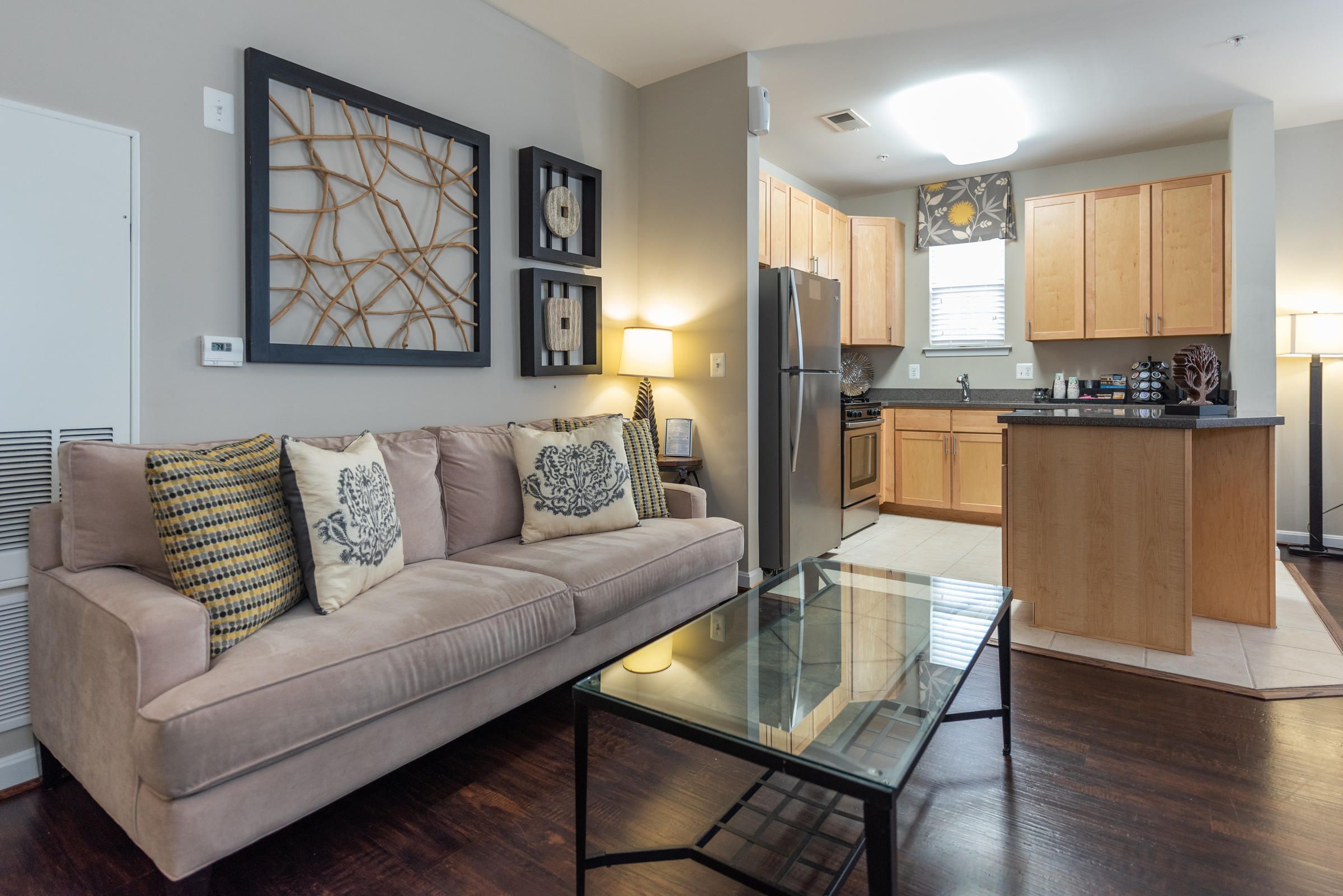 Modern living room with beige sofa, glass coffee table, and open kitchen with wooden cabinets in the background.