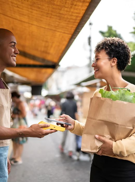 Smiling woman pays vendor at outdoor market, holding grocery bag filled with fresh produce.