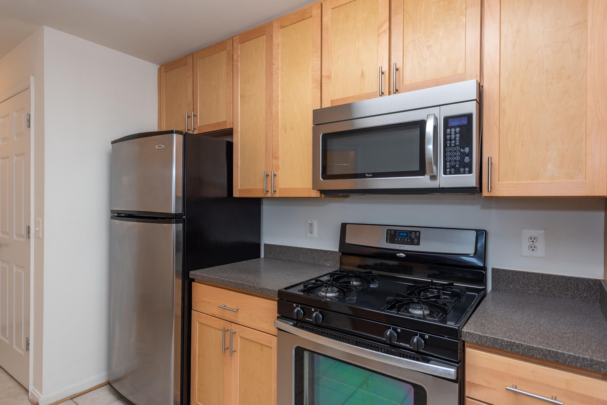 Modern kitchen with light wood cabinets, stainless steel fridge, microwave, and gas stove with black countertops.