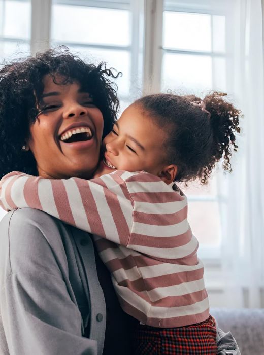 Smiling woman holding a laughing young girl who hugs her, both happy in a bright room with large windows.